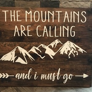 Puede incluir: Un letrero de madera r&uacute;stico con el texto "The Mountains Are Calling - and I Must Go" pintado en blanco. El letrero tambi&eacute;n presenta una silueta blanca de una cordillera.
