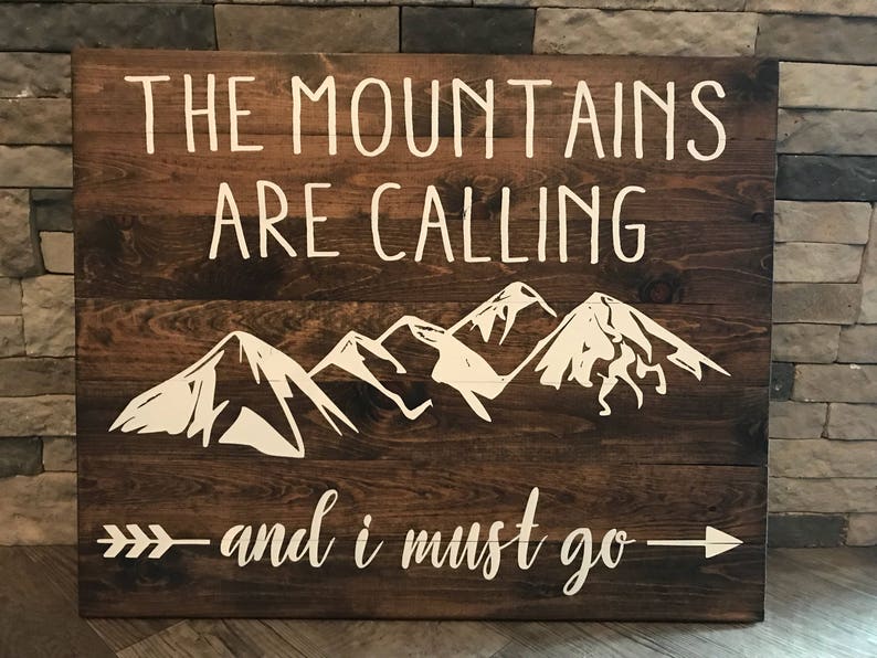 Puede incluir: Un letrero de madera r&uacute;stico con el texto "The Mountains Are Calling - and I Must Go" pintado en blanco. El letrero tambi&eacute;n presenta una silueta blanca de una cordillera.