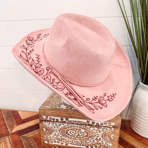 May include: A pink suede cowboy hat with a red floral design. The hat has a brown leather band and a tassel.