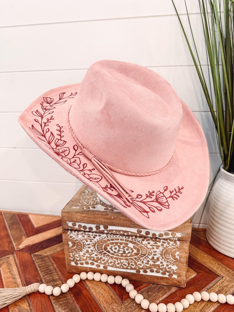 May include: A pink suede cowboy hat with a red floral design. The hat has a brown leather band and a tassel.