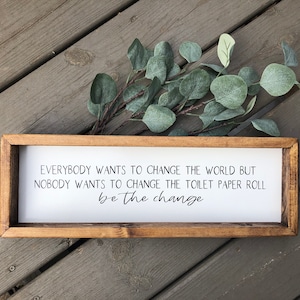 May include: A wooden framed sign with a white background and black text that reads "Everybody wants to change the world but nobody wants to change the toilet paper roll be the change".