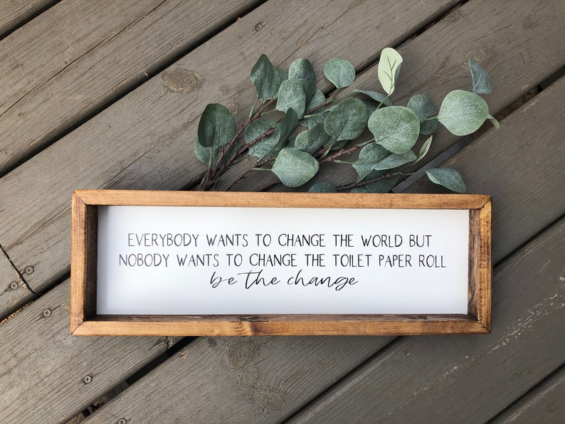 May include: A wooden framed sign with a white background and black text that reads "Everybody wants to change the world but nobody wants to change the toilet paper roll be the change".