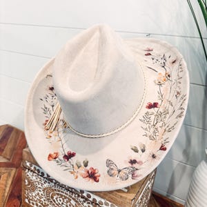 May include: A light beige cowboy hat with a floral and butterfly design. The hat features a cream-colored brim with a decorative floral pattern in shades of red, orange, and green. A braided trim and tassel add detail.