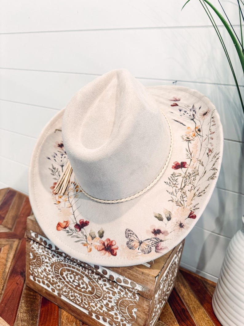 May include: A light beige cowboy hat with a floral and butterfly design. The hat features a cream-colored brim with a decorative floral pattern in shades of red, orange, and green. A braided trim and tassel add detail.