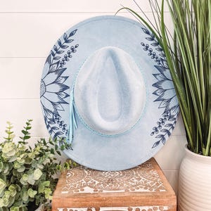 May include: A light blue felt hat with a blue floral design. The hat has a light blue tassel and a wide brim.