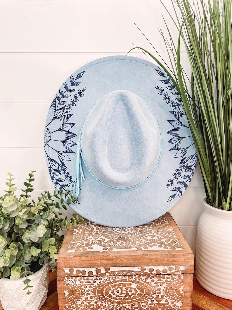 May include: A light blue felt hat with a blue floral design. The hat has a light blue tassel and a wide brim.