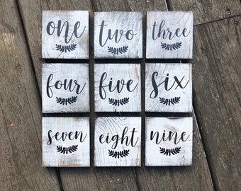 May include: Nine white wooden blocks with black painted numbers and a simple leaf design. The blocks are arranged in three rows of three, with the numbers one through nine.