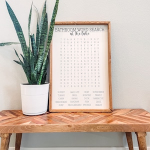 May include: A wooden sign with a bathroom word search puzzle on it. The puzzle is themed around a lake and includes words like "sunset", "lake life", "boat", "sunscreen", "tubing", "grill", "bonfire", "family", "cabin", "kayak", "dock", "barbecue", "wakeboard", "sandbar", "fish", "lake", "surf", "paddleboard", "smores", and "beach".