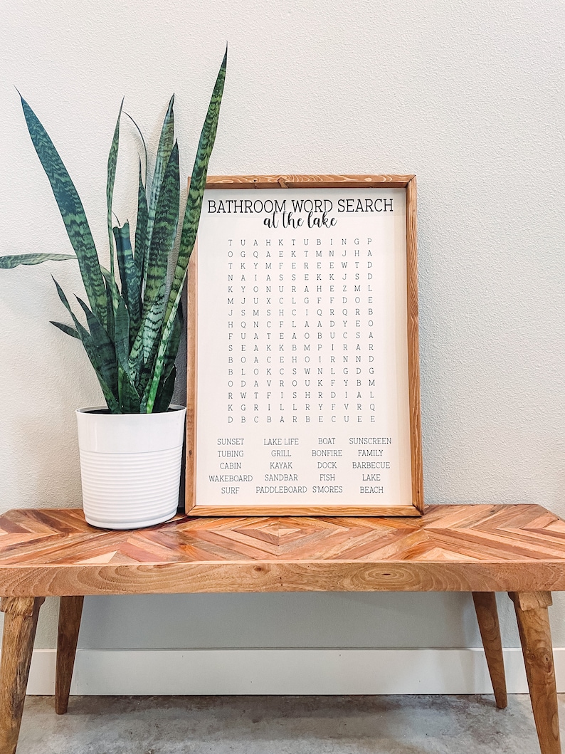 May include: A wooden sign with a bathroom word search puzzle on it. The puzzle is themed around a lake and includes words like "sunset", "lake life", "boat", "sunscreen", "tubing", "grill", "bonfire", "family", "cabin", "kayak", "dock", "barbecue", "wakeboard", "sandbar", "fish", "lake", "surf", "paddleboard", "smores", and "beach".