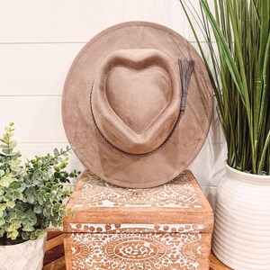 May include: A tan-colored hat with a heart-shaped crown and a decorative tassel. The hat is displayed on a wooden box with white carved designs. The box sits on a wooden table with a geometric pattern. Green plants in white pots are in the background.