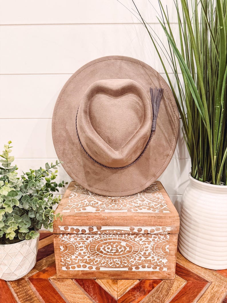 May include: A tan-colored hat with a heart-shaped crown and a decorative tassel. The hat is displayed on a wooden box with white carved designs. The box sits on a wooden table with a geometric pattern. Green plants in white pots are in the background.