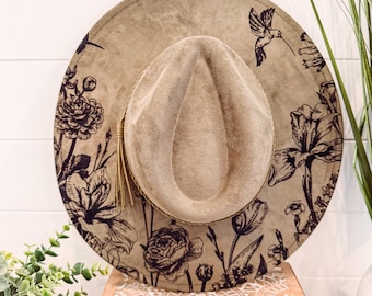 May include: A tan-colored fedora hat with a wide brim, adorned with a black floral and hummingbird design. The hat features a matching tan crown and a decorative tassel. The hat is displayed on a wooden surface.