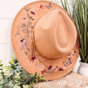 Op de afbeelding: Een perzikkleurige fedora-hoed met een bloemen- en vlinderontwerp. De hoed heeft een decoratieve kwast en een bruine band. De rand is versierd met een delicaat patroon van bloemen en vlinders.