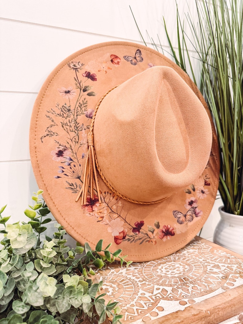 Op de afbeelding: Een perzikkleurige fedora-hoed met een bloemen- en vlinderontwerp. De hoed heeft een decoratieve kwast en een bruine band. De rand is versierd met een delicaat patroon van bloemen en vlinders.