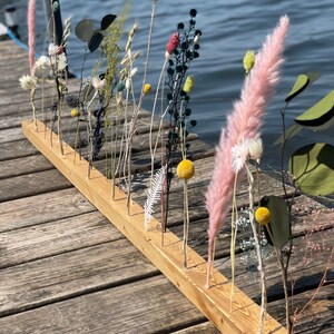 Pu&ograve; includere: Un asse di legno con pi&ugrave; fori praticati, utilizzato per tenere fiori ed erbe secche. L'asse &egrave; appoggiata su una superficie di legno. I fiori includono erba della pampa secca, respiro di bambino secco, eucalipto secco e pompon gialli secchi.