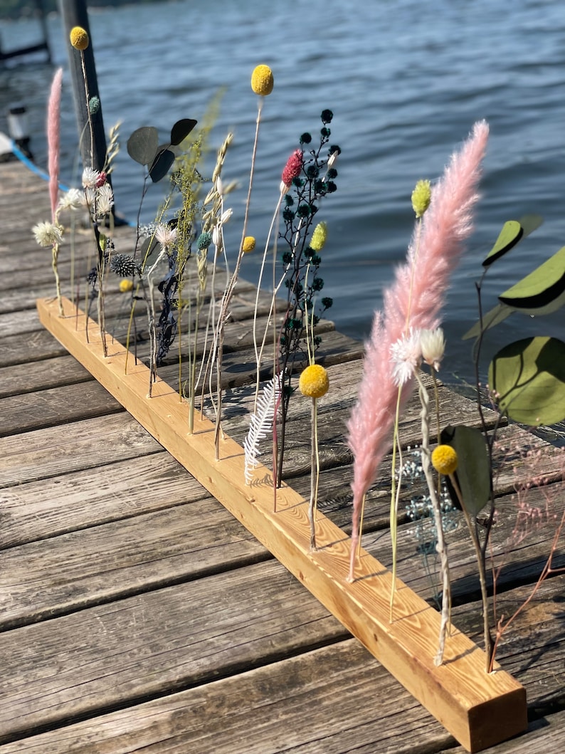 Pu&ograve; includere: Un asse di legno con pi&ugrave; fori praticati, utilizzato per tenere fiori ed erbe secche. L'asse &egrave; appoggiata su una superficie di legno. I fiori includono erba della pampa secca, respiro di bambino secco, eucalipto secco e pompon gialli secchi.