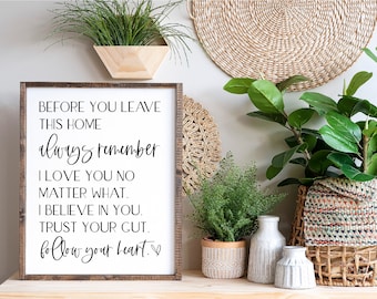 May include: A framed sign with a white background and black text that reads "Before you leave this home always remember I love you no matter what. I believe in you. Trust your gut. Follow your heart." The sign is hanging on a wall with plants and a woven mat.