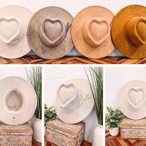 May include: A collection of wide-brimmed hats in various neutral colors, including cream, taupe, and brown. Each hat features a heart-shaped crown and a decorative band. The hats are displayed on a wooden surface and decorative boxes, showcasing a bohemian style.