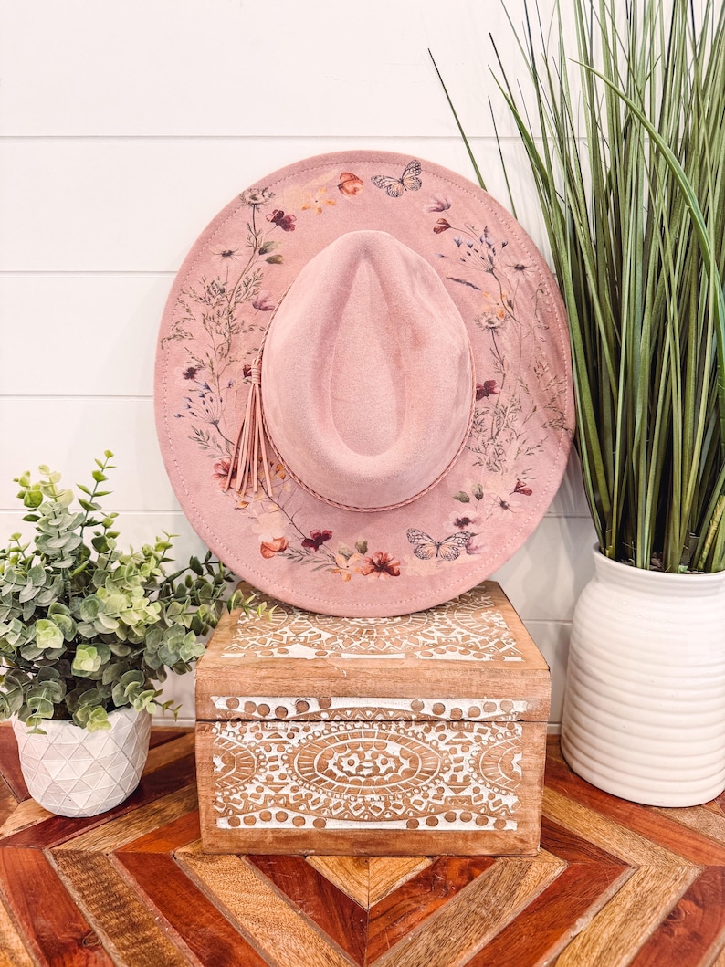 May include: A pink suede hat with a floral and butterfly print. The hat has a wide brim and a tassel.