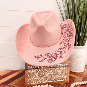 May include: A pink cowboy hat with a floral design in maroon. The hat is sitting on a wooden box with a decorative pattern.