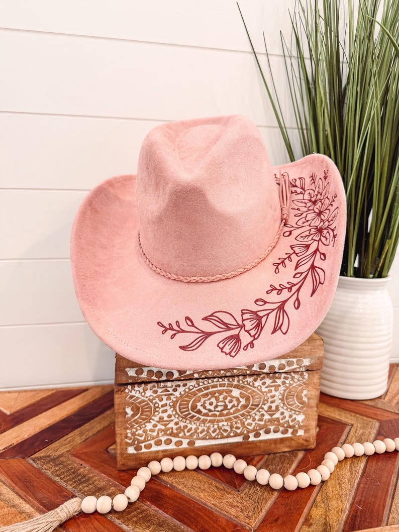 May include: A pink cowboy hat with a floral design in maroon. The hat is sitting on a wooden box with a decorative pattern.