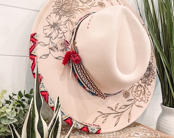 May include: A cream-colored wide-brimmed hat with a floral design and red and blue trim. The hat has a feather accent and a tassel.