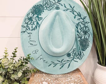 May include: A light blue felt fedora hat with a floral design and the text "wild & free" in a cursive font.