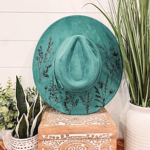 May include: A teal blue suede hat with a black floral design. The hat is sitting on a wooden box with a carved design.