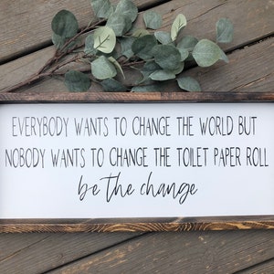 May include: A wooden sign with the text "EVERYBODY WANTS TO CHANGE THE WORLD BUT NOBODY WANTS TO CHANGE THE TOILET PAPER ROLL be the change".
