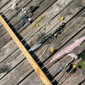 Pu&ograve; includere: Un bastone di legno con fiori e foglie secche attaccati. Il bastone &egrave; appoggiato su una superficie di legno. I fiori sono in tonalit&agrave; di rosa, bianco e giallo. Il fogliame &egrave; verde.