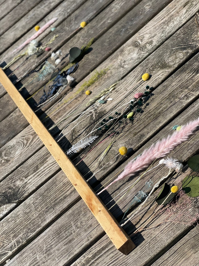 Pu&ograve; includere: Un bastone di legno con fiori e foglie secche attaccati. Il bastone &egrave; appoggiato su una superficie di legno. I fiori sono in tonalit&agrave; di rosa, bianco e giallo. Il fogliame &egrave; verde.