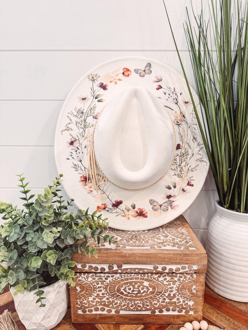 May include: A white wide-brimmed hat with a floral and butterfly design. The hat has a brown leather band and a brown tassel. The hat is sitting on a wooden box with a carved design.