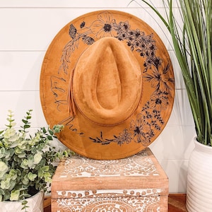 May include: A brown suede hat with a black floral design and the word "Lilac" written on the brim.
