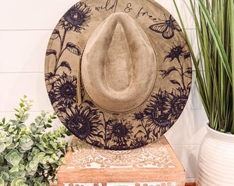 Pode incluir: Um chapéu fedora bege com aba larga, com um design de girassóis e borboletas em tinta preta. O chapéu tem as palavras "wild & free" em escrita. Uma borla dourada pende do chapéu. O chapéu é exibido em uma caixa de madeira decorativa.