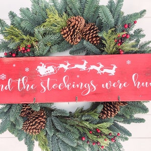 Puede incluir: Un letrero de madera rojo con texto blanco que dice "and the stockings were hung". El letrero tiene una silueta blanca de Santa Claus en un trineo tirado por renos. El letrero está decorado con copos de nieve y está colgado de una corona de Navidad verde.
