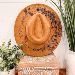 Puede incluir: Un sombrero de gamuza marr&oacute;n con un dise&ntilde;o floral negro y la palabra "Lilac" escrita en el borde.