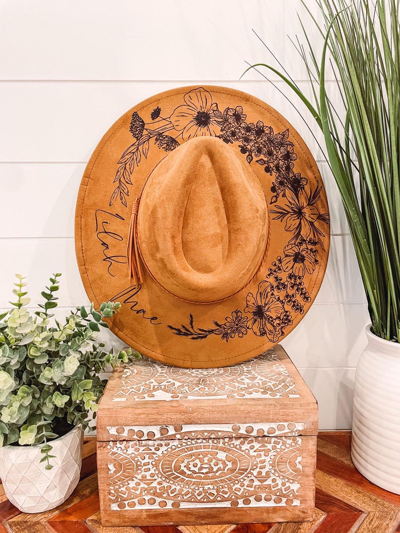 Puede incluir: Un sombrero de gamuza marr&oacute;n con un dise&ntilde;o floral negro y la palabra "Lilac" escrita en el borde.