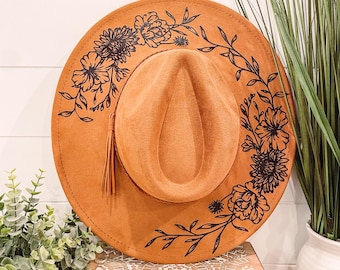 May include: A brown suede hat with a black floral design around the brim. The hat has a brown suede band and a brown tassel.