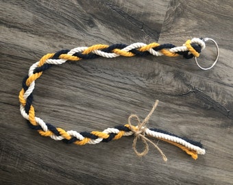 May include: Braided rope keychain in navy blue, white, and gold. The rope is tied with a natural twine bow. A silver metal ring is attached to the end of the rope.