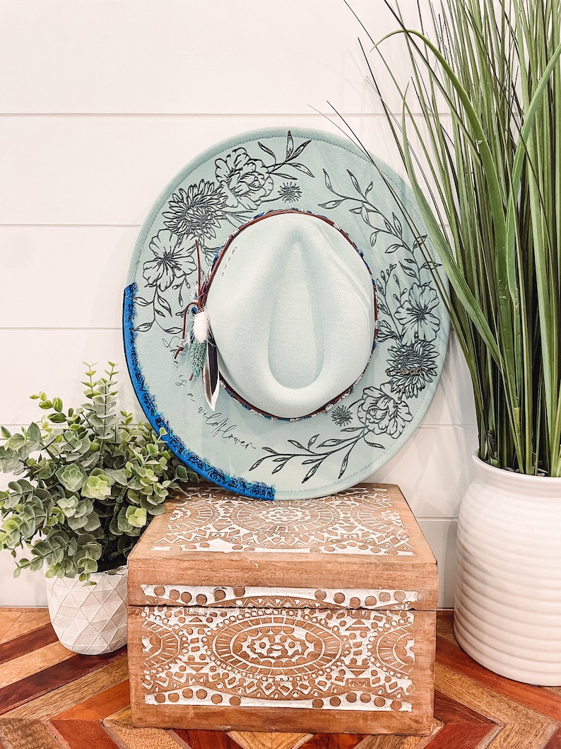 May include: A light blue hat with a floral design and the text "It is a wildflower" on the brim. The hat is sitting on a wooden box with a carved design. A potted plant is in the background.