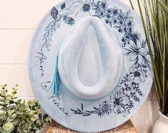 May include: A pale blue fedora hat featuring a floral pattern and the text "Amore la Femme" in black. The hat has a turquoise band and tassel. The hat is displayed on a wooden box.