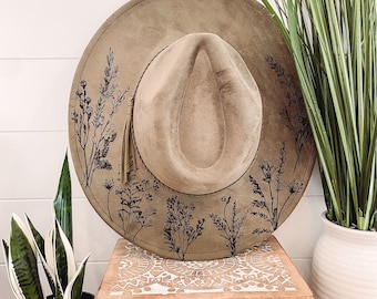 May include: A brown suede hat with a wide brim and a black floral design. The hat is sitting on top of a wooden box with an intricate carved design.