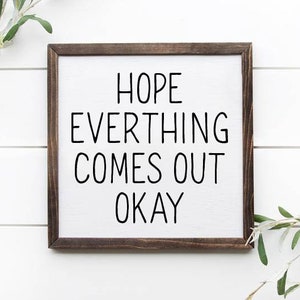 Puede incluir: Un letrero enmarcado en madera con el texto "HOPE EVERYTHING COMES OUT OKAY" en letras negras sobre fondo blanco.