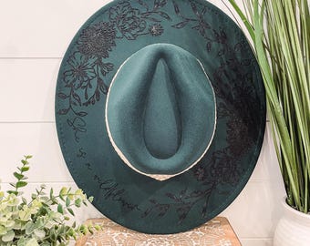 May include: A dark green fedora hat with a wide brim, featuring black floral designs and the text "She is a wildflower." The hat has a cream-colored band and is displayed on a wooden box.