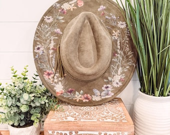 May include: A wide-brimmed olive green hat with a floral design and a tassel. The hat is decorated with pink, purple, and white flowers and butterflies. The hat is resting on a wooden box with a white carved design.