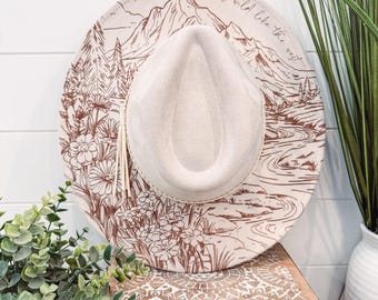 Pode incluir: Um chapéu de feltro bege com um desenho de montanha e flores marrons. O chapéu tem uma aba larga e uma faixa de couro marrom com o texto "She's wild like the west" escrito nele.