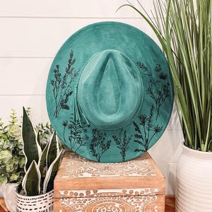 Puede incluir: Un sombrero de gamuza azul turquesa con un dise&ntilde;o floral negro. El sombrero est&aacute; sentado en una caja de madera con un dise&ntilde;o tallado.