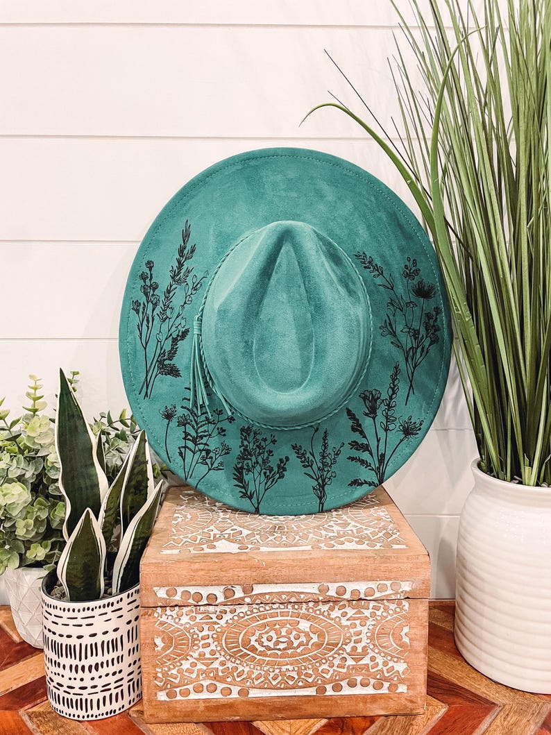Puede incluir: Un sombrero de gamuza azul turquesa con un dise&ntilde;o floral negro. El sombrero est&aacute; sentado en una caja de madera con un dise&ntilde;o tallado.