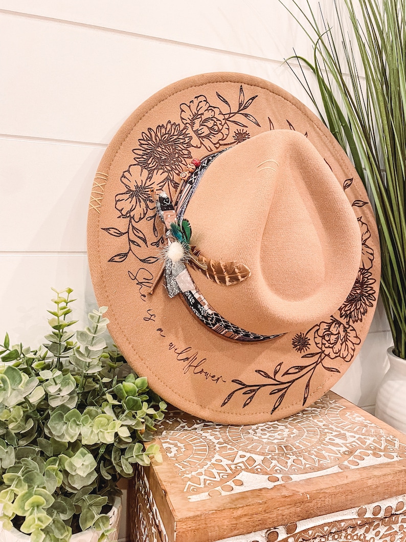 May include: A brown felt fedora hat with a floral design and the text "She is a wildflower" engraved on the side. The hat has a black and white patterned band with a feather accent.