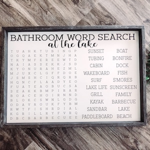 Puede incluir: Un letrero de madera blanco con marco negro y el texto "Bathroom Word Search at the Lake" impreso en negro. El letrero presenta un crucigrama de palabras con palabras relacionadas con el lago, como "barco", "caba&ntilde;a", "pescado", "parrilla", "kayak", "lago", "tabla de remo", "atardecer", "surf", "tubing" y "wakeboard".